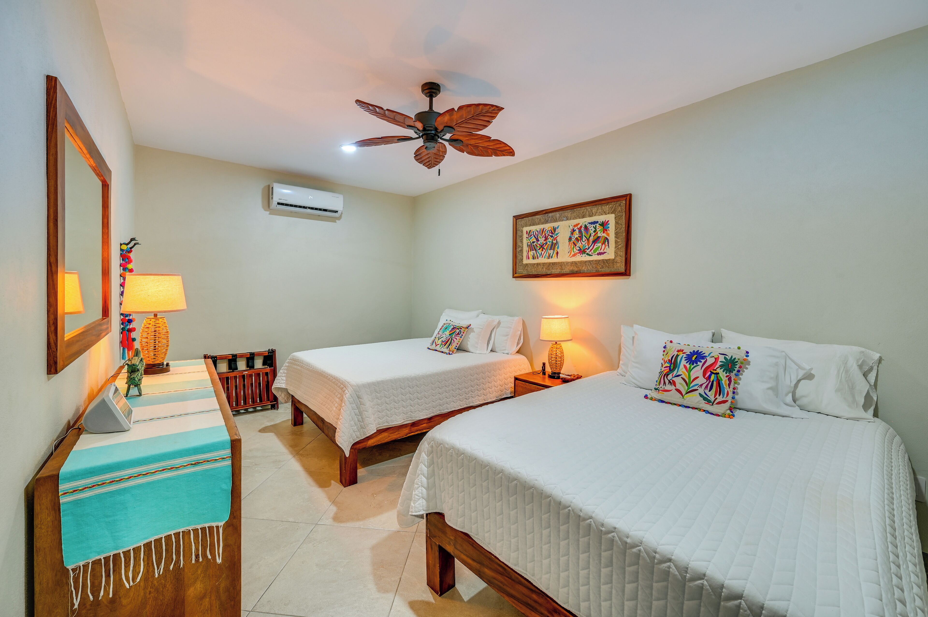 A hotel room with two beds, a desk with a lamp, a ceiling fan, an air conditioning unit, and a framed artwork on the wall.
