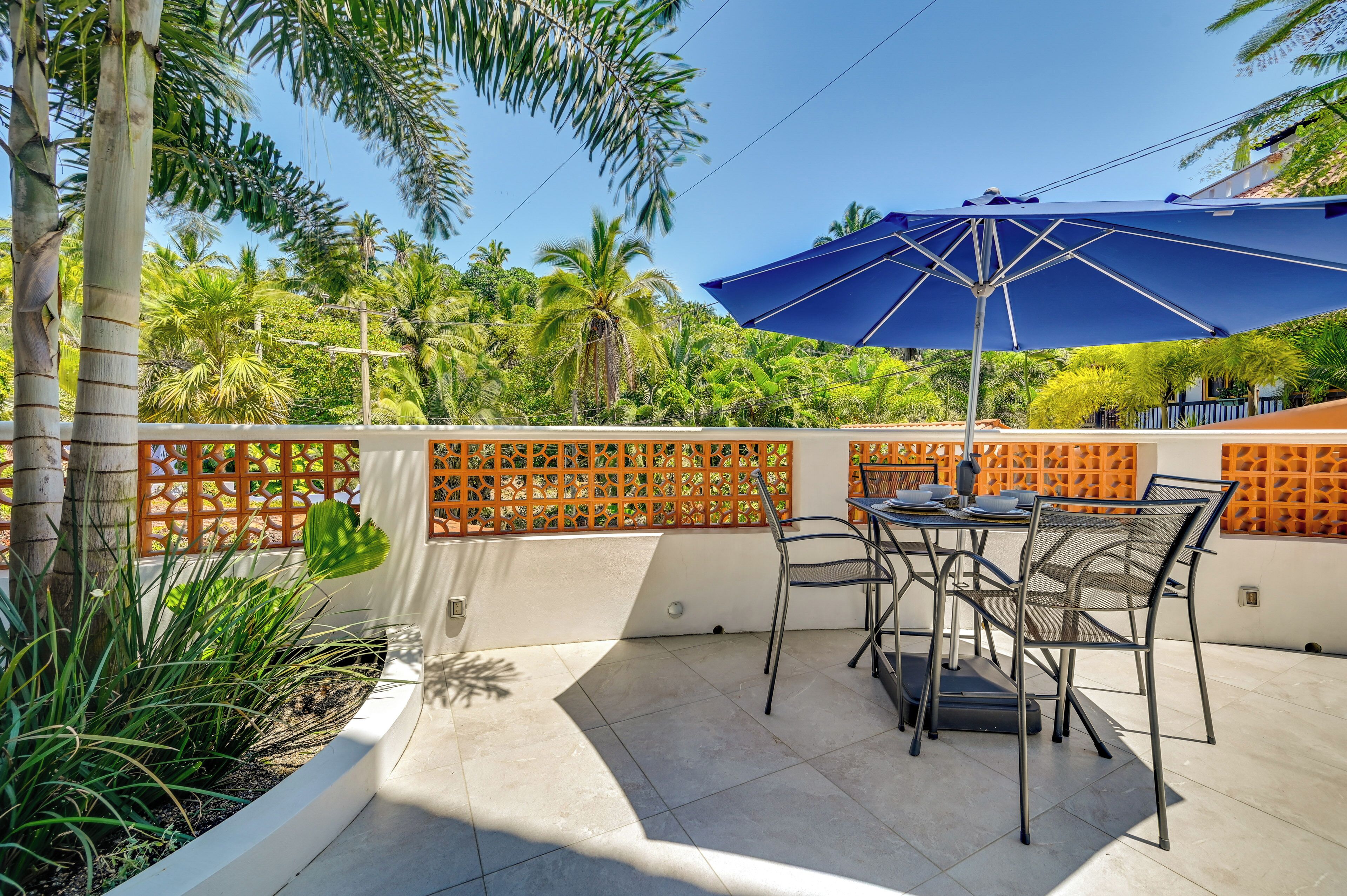 Un patio al aire libre con una mesa, sillas, un paraguas azul y una cerca decorativa.