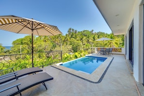 A rooftop pool with a view of the ocean, surrounded by greenery and a dining area with chairs and tables.