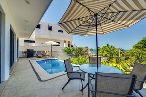 A modern outdoor pool area with a table and chairs, a large umbrella, and a view of the ocean.