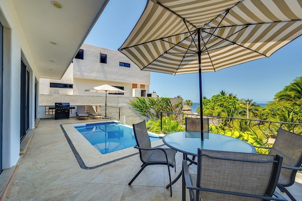 A modern outdoor pool area with a table and chairs, a large umbrella, and a view of the ocean.
