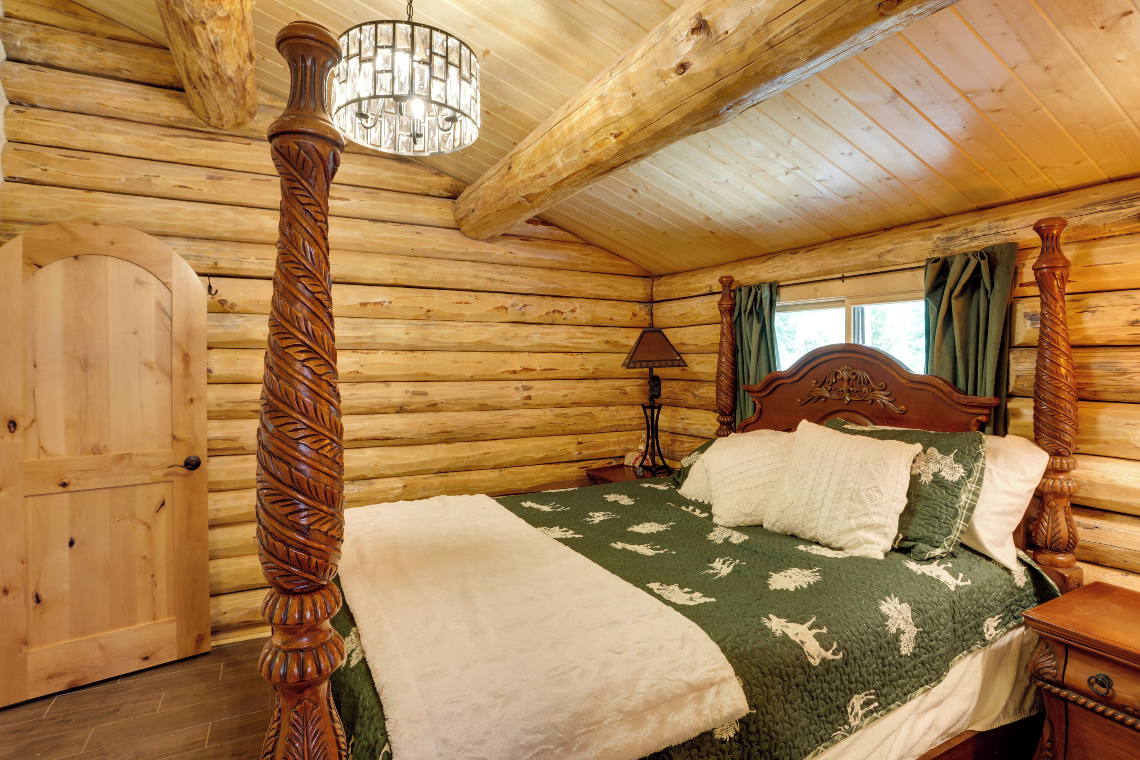 A log cabin bedroom with a four-poster bed, wooden walls, a chandelier, and a window with curtains.