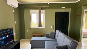 A living room with a sectional sofa, a flat-screen TV, a window with curtains, a door marked 'EXIT', and a bench.