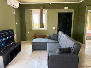 A living room with a sectional sofa, a flat-screen TV, a window with curtains, a door marked 'EXIT', and a bench. - Artemida Olive Gem - Sunny Balcony Views Parking (Spata-Artemida)