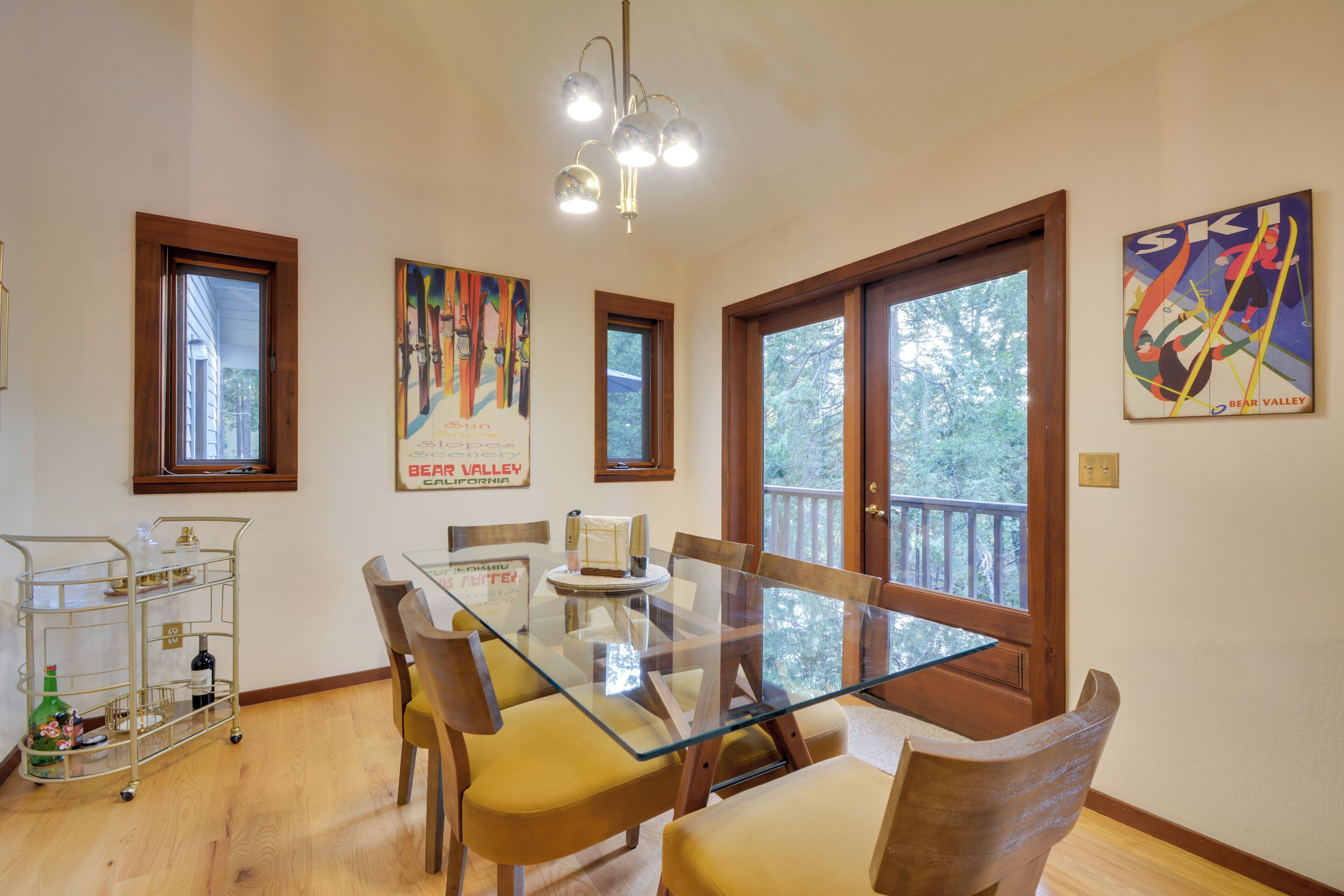 A dining area with a glass table, wooden chairs, a metal cart, and a window with a view of trees.