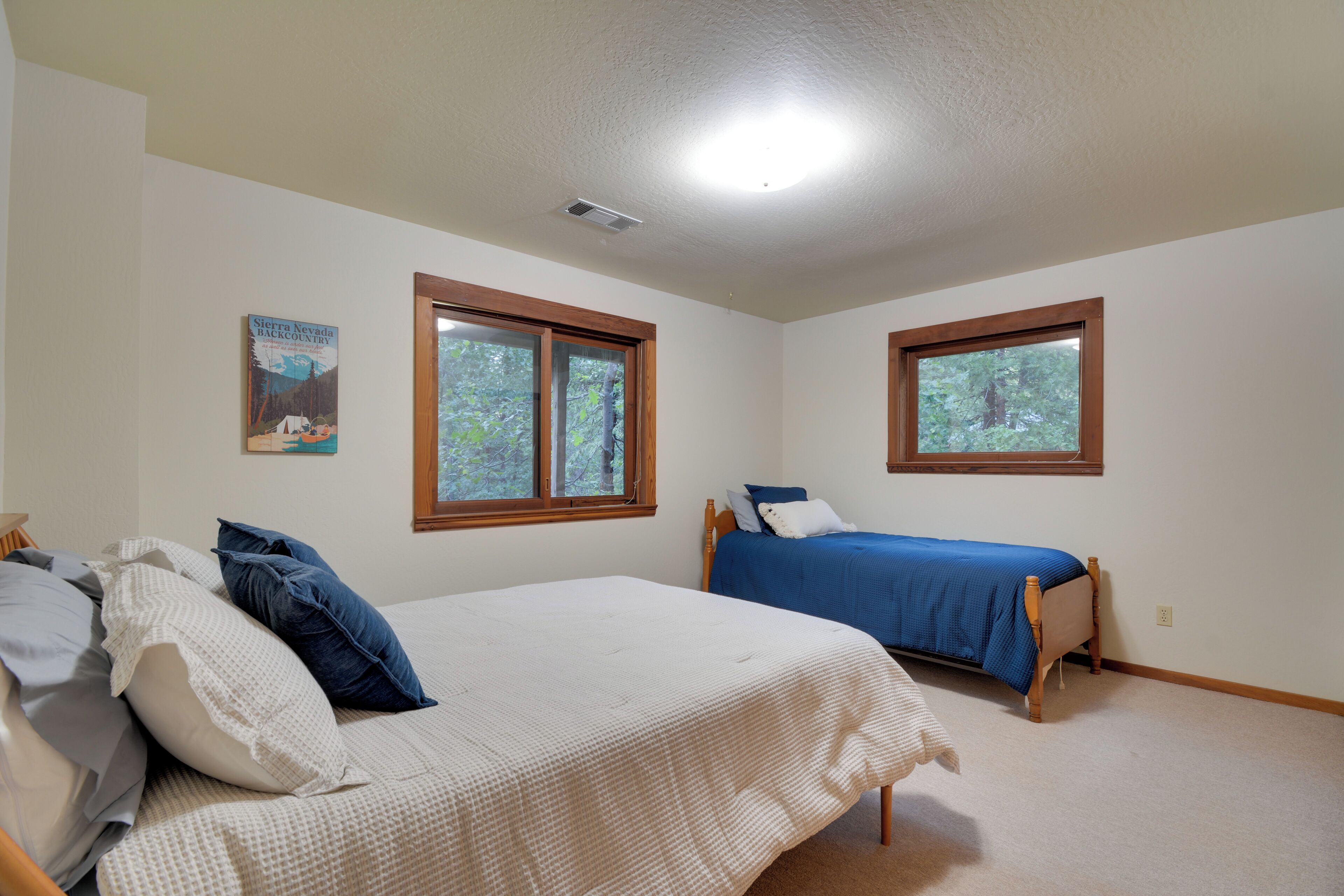 A bedroom with two beds, a ceiling light, and two windows with views of trees.
