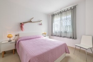 Un dormitorio con cama, mesita de noche con lámparas, una silla, ventana con cortinas y un adorno en la pared.