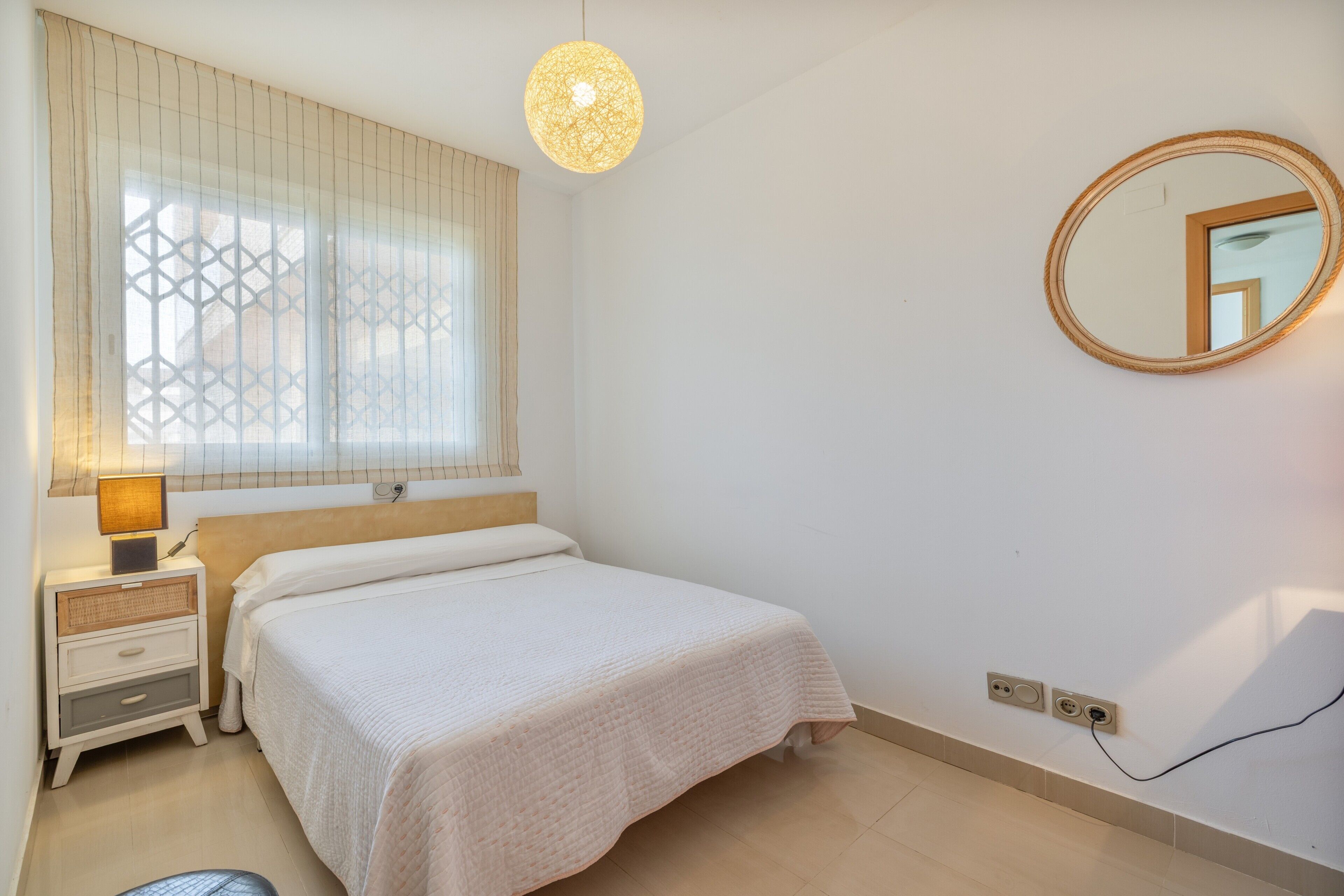 A bedroom with a bed, bedside lamp, window with blinds, and a wall-mounted mirror.