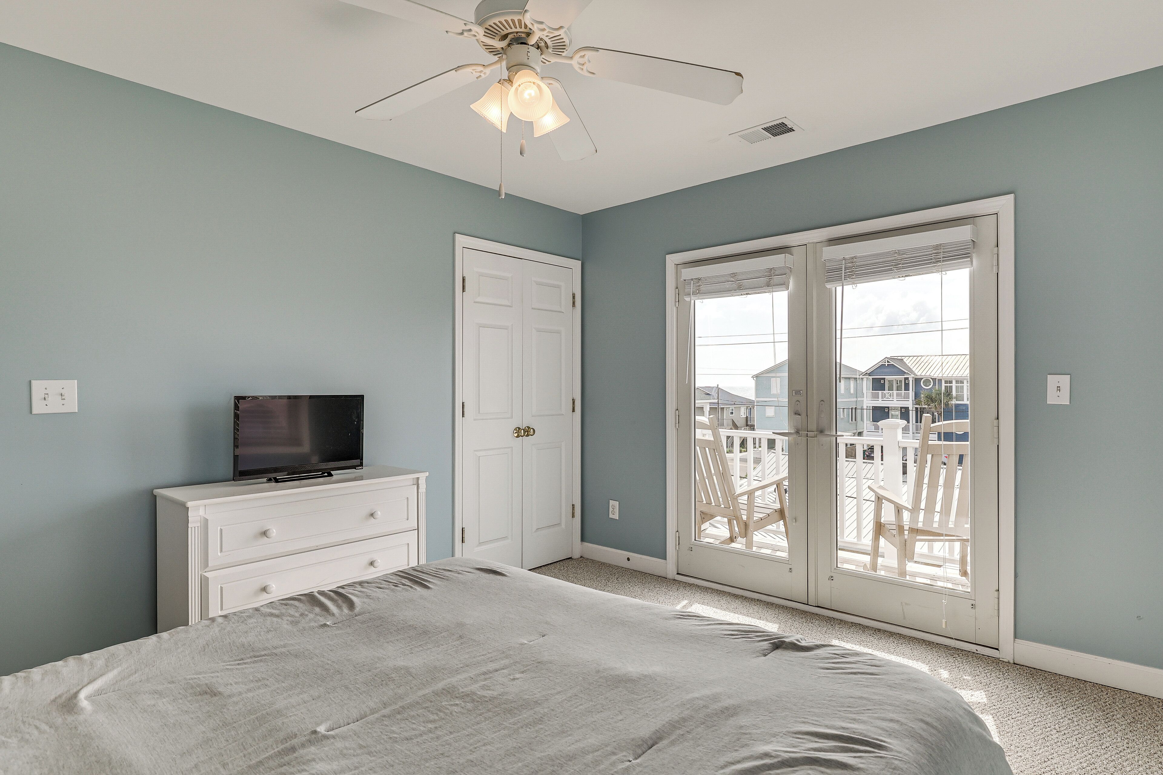 A bedroom with a bed, a dresser, a television, a ceiling fan, and a sliding glass door leading to a balcony.