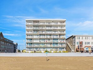 Ein mehrstöckiges Hotel mit Balkonen, feinem Sand vor der Fassade und blauem, wolkenlosem Himmel.