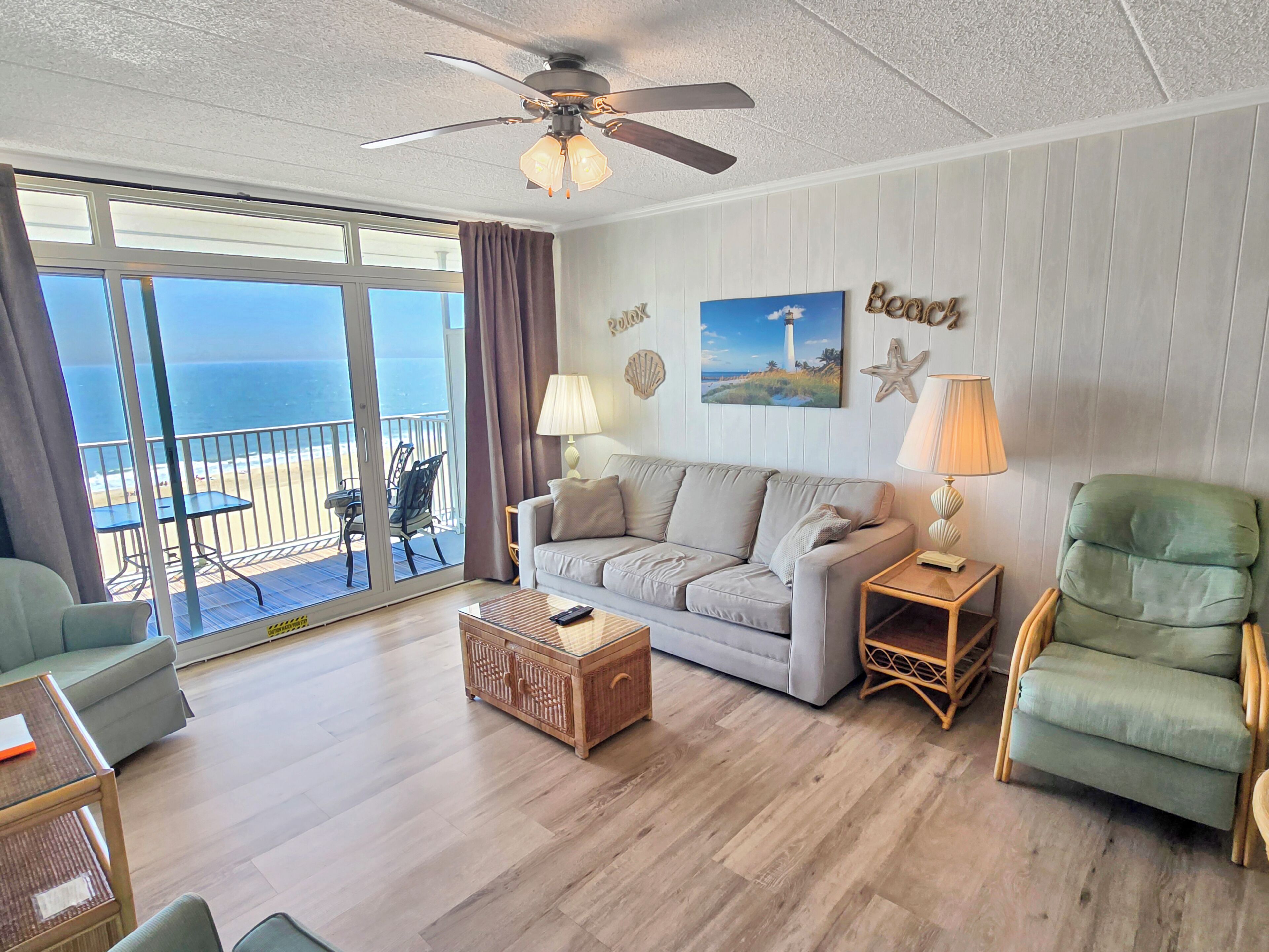 A living room with a sofa, armchairs, a coffee table, and a ceiling fan. There is a balcony with a view of the ocean.