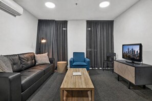 Ein modernes Hotelzimmer mit einer Ledersofa, einem blauen Sessel, einem Holztisch und einem kleinen runden Beistelltisch sowie einem Fernseher.