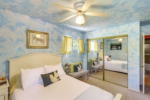Um quarto com cama, um banquinho, um ventilador de teto, um espelho e uma porta.