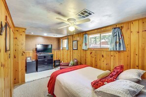 Quarto com paredes de madeira, cama com várias almofadas, ventoinha no teto, televisão e uma pequena escrivaninha com gavetas.