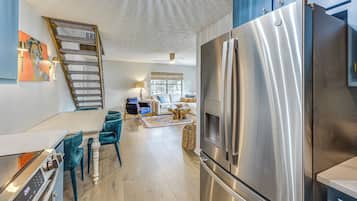 A modern kitchen with a stainless steel refrigerator, a dining area with teal chairs, and a staircase leading to an upper level.