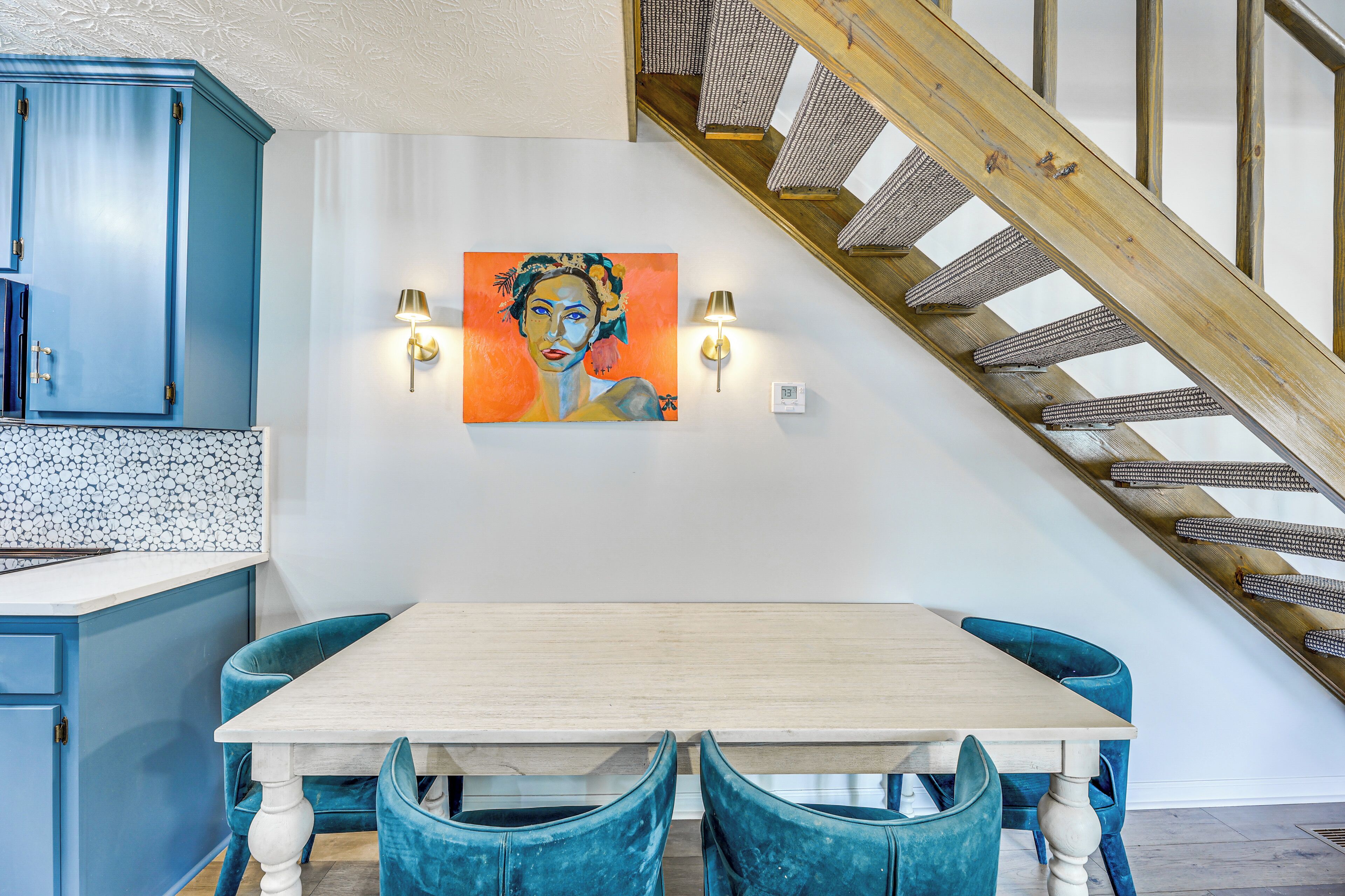 Un comedor con mesa de madera y sillas tapizadas en azul, un cuadro en la pared y una escalera con barandillas de madera.