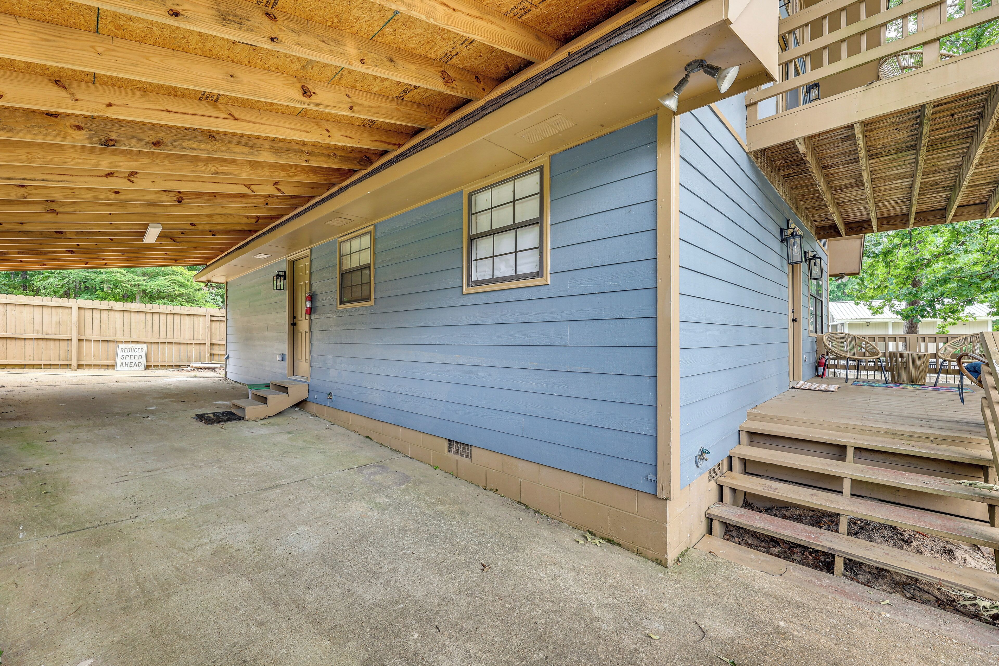 A covered outdoor area with a wooden deck, a blue building, and a fenced yard.