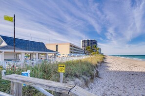 Ett hotell vid stranden med en skylt som uppmanar besökare att hålla sig borta från sanddynerna, ett trästaket och utsikt över havet och byggnaderna i bakgrunden.