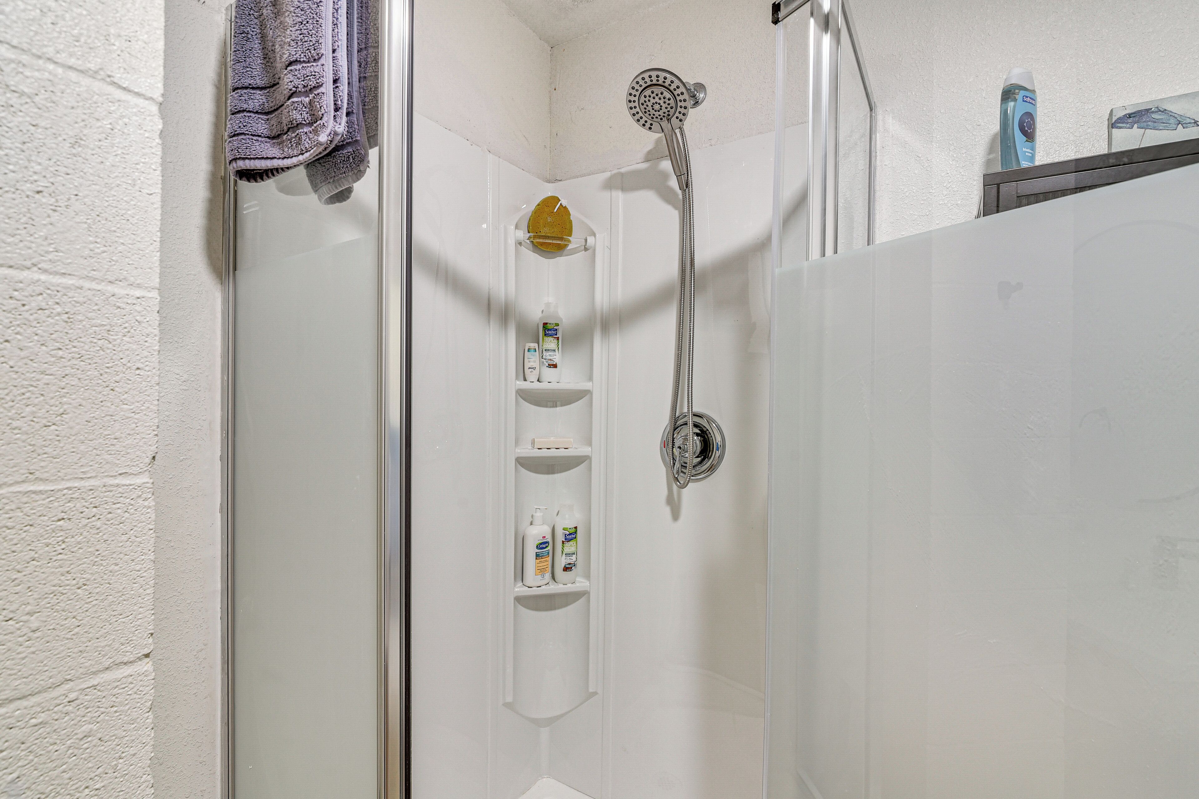 Una ducha con cabezal de ducha de mano, puerta de vidrio y estante con artículos de tocador.