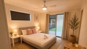 Ein Schlafzimmer mit einem Bett, Nachttischen, einem Deckenventilator, einem Fenster mit Jalousien, einer Glastür, einem Blumentopf, einem kleinen Tisch mit einer Kerze und einem Sessel mit einem gestreiften Kissen.