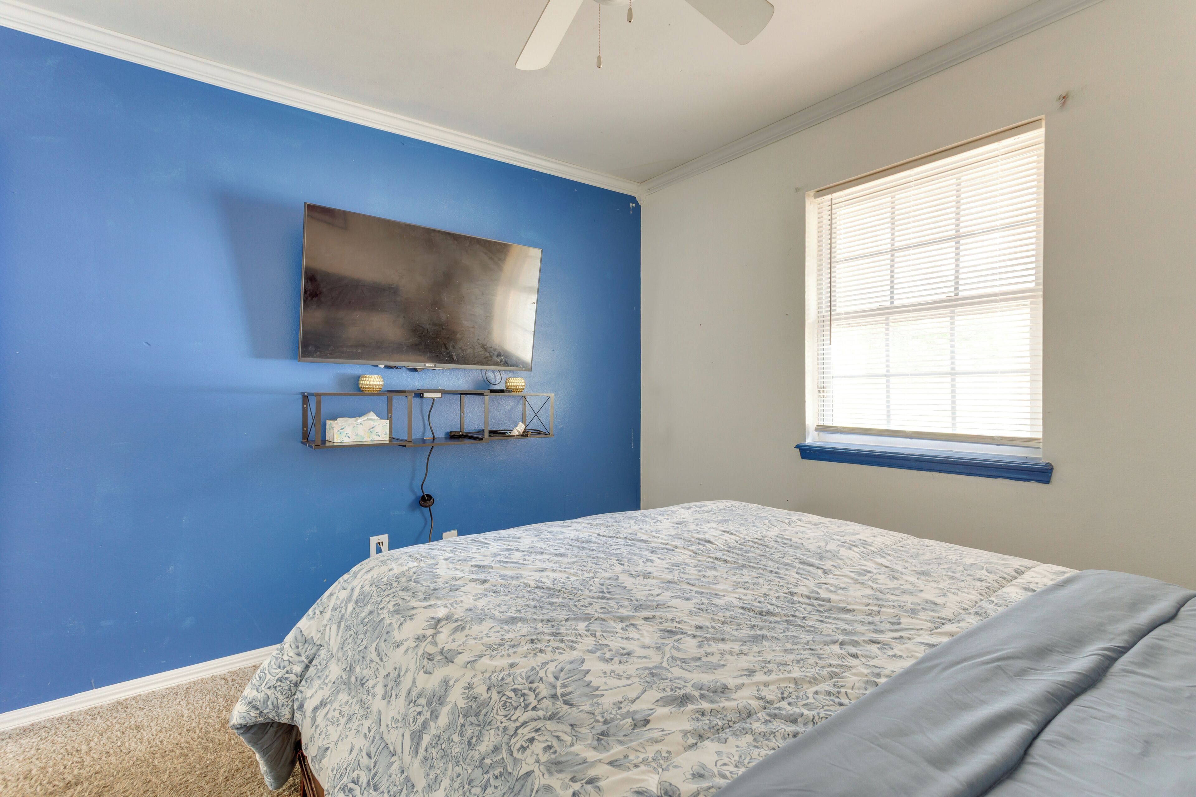 Une chambre avec un mur bleu, un lit recouvert d’une couvre-lit à motifs floraux, un téléviseur fixé au mur et une fenêtre avec des stores.