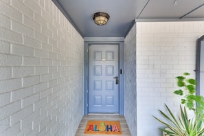 Un couloir avec une porte bleue, un paillasson orné du mot « ALOHA » et d’un ananas, et une plante en pot.