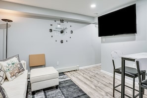 A modern living room with a wall clock, a flat-screen TV, a sofa, a coffee table, and a dining area with a table and chairs. - Work & Play in Baldwin! Cozy Apartment w/ Grill (Baldwin)