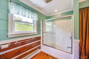 A bathroom with a shower, a window with a curtain, wooden paneling, and a view of a grassy area.