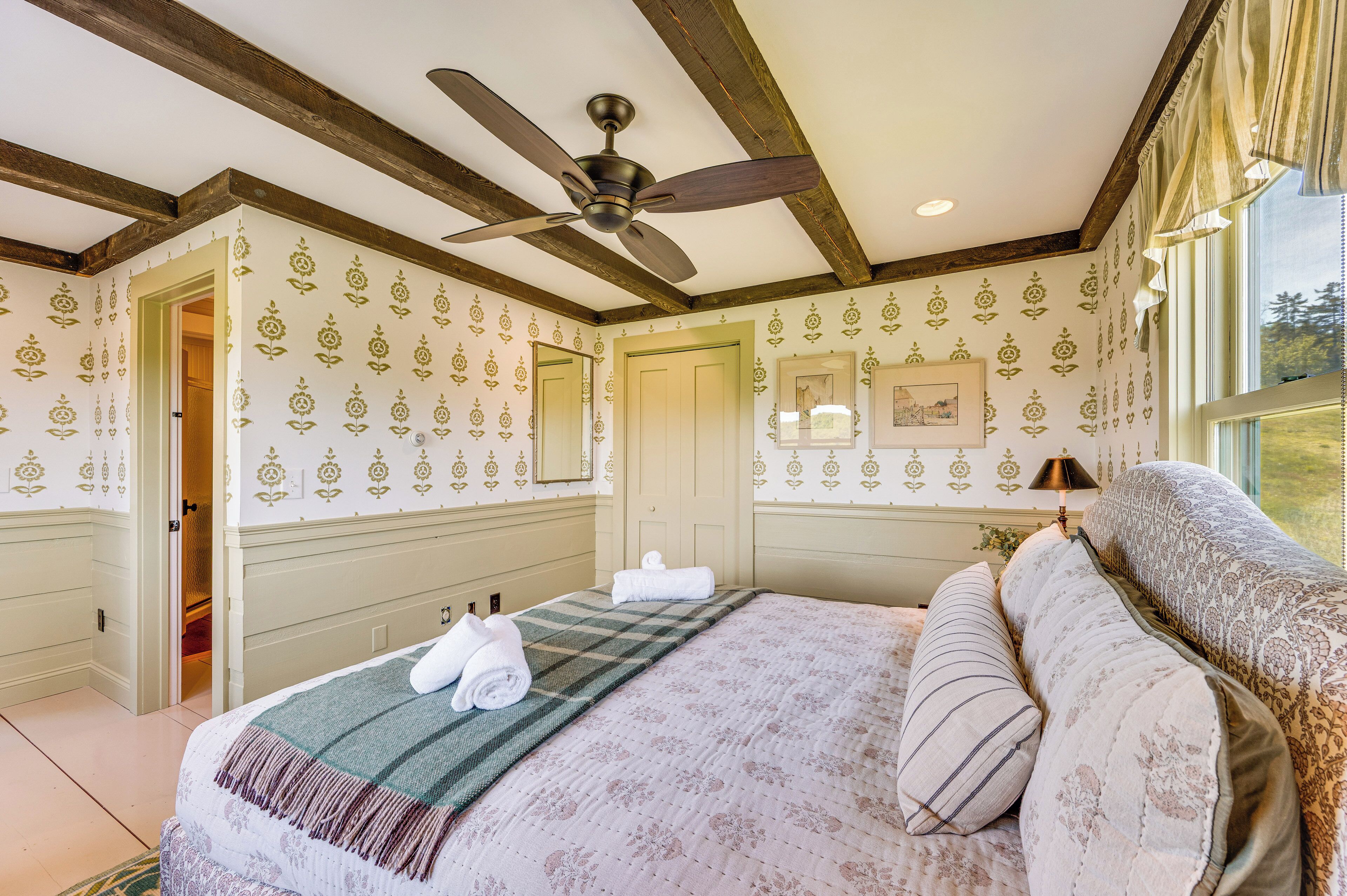 A bedroom with a bed, a ceiling fan, a window with curtains, and a door leading to another room.