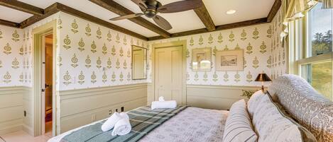 Une chambre à coucher comprenant un lit, un ventilateur de plafond, une fenêtre avec des rideaux et une porte donnant sur une autre pièce.