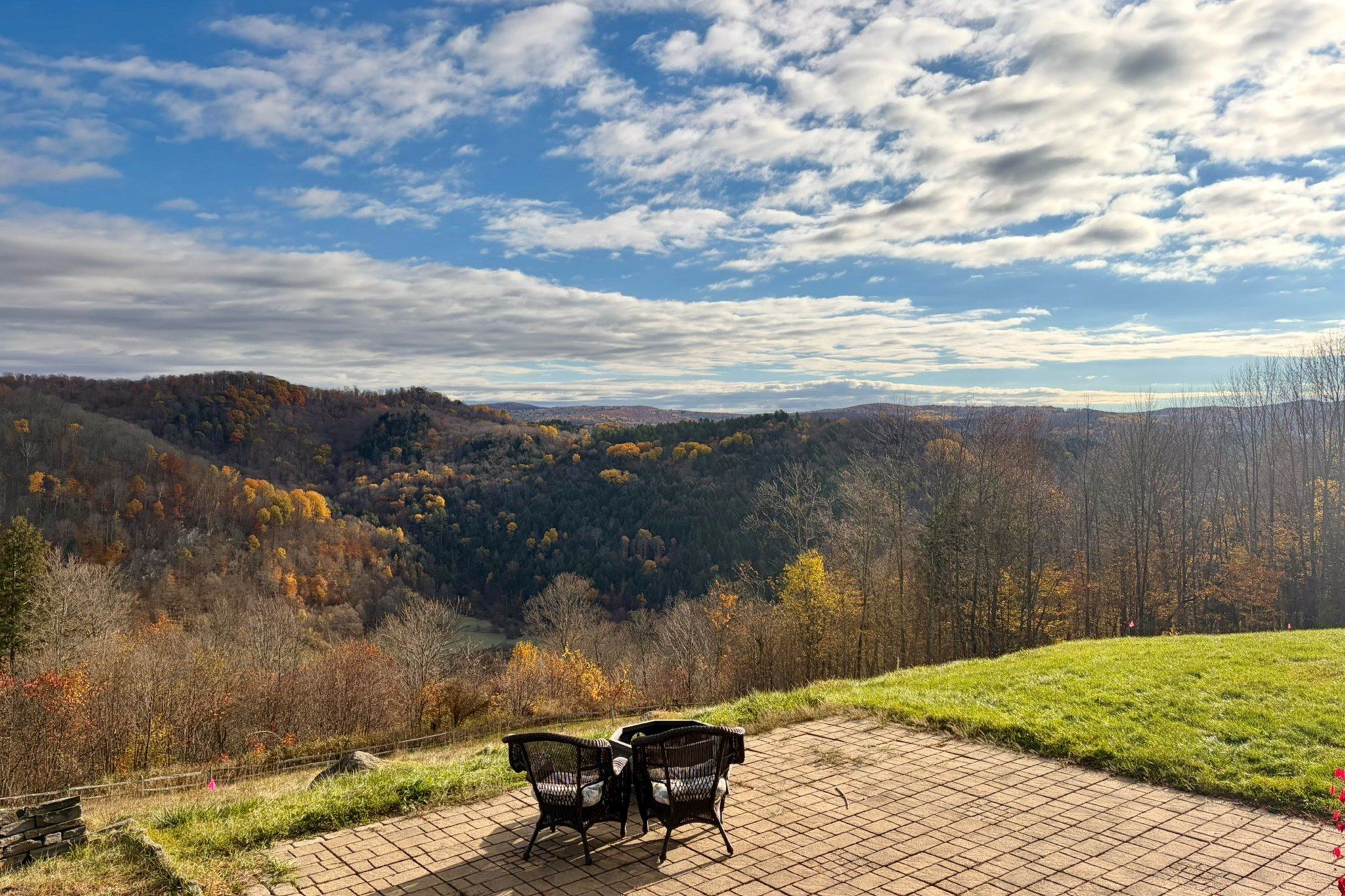 A paved outdoor seating area with two chairs and a table, overlooking a forested valley with autumn foliage.