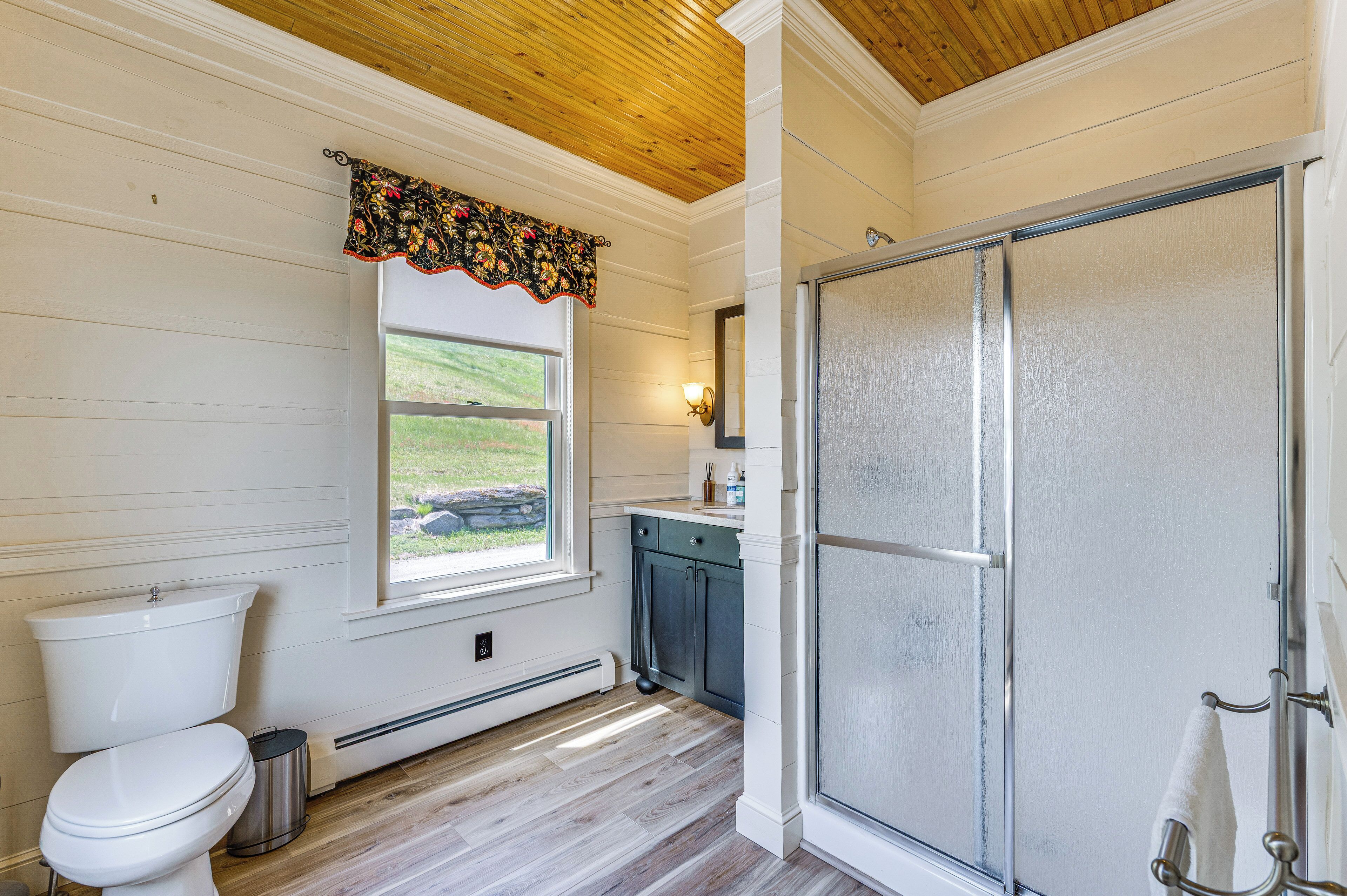 Een badkamer met toilet, een raam met bloemen gordijn, douche met een mat glazen deur en een wastafel met kastjes.