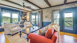 A room with a sofa, armchair, and a coffee table. There is a bookshelf, a window with curtains, and a door with a view. - 95-acre Countryside Estate! Luxe Vermont Farmhouse (Chelsea)
