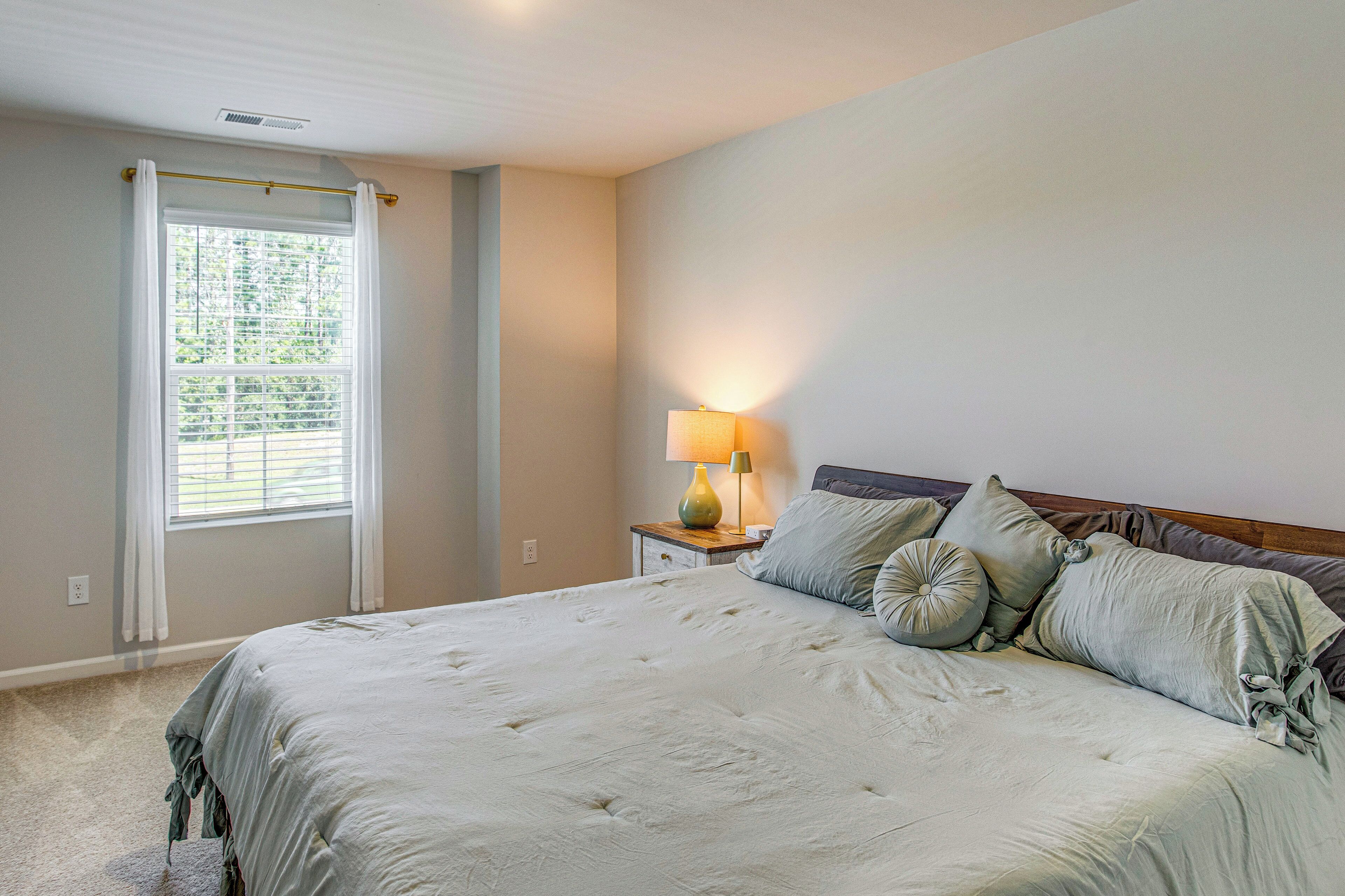 Une chambre à coucher avec un lit, une table de chevet avec une lampe, une fenêtre avec des rideaux et une vue sur la verdure à l’extérieur.