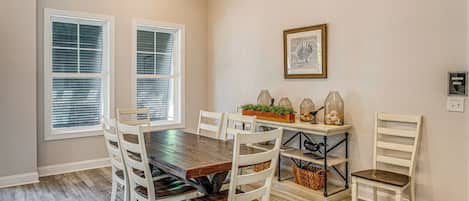 Comedor con mesa de madera, sillas blancas, aparador con adornos y dos ventanas con persianas.