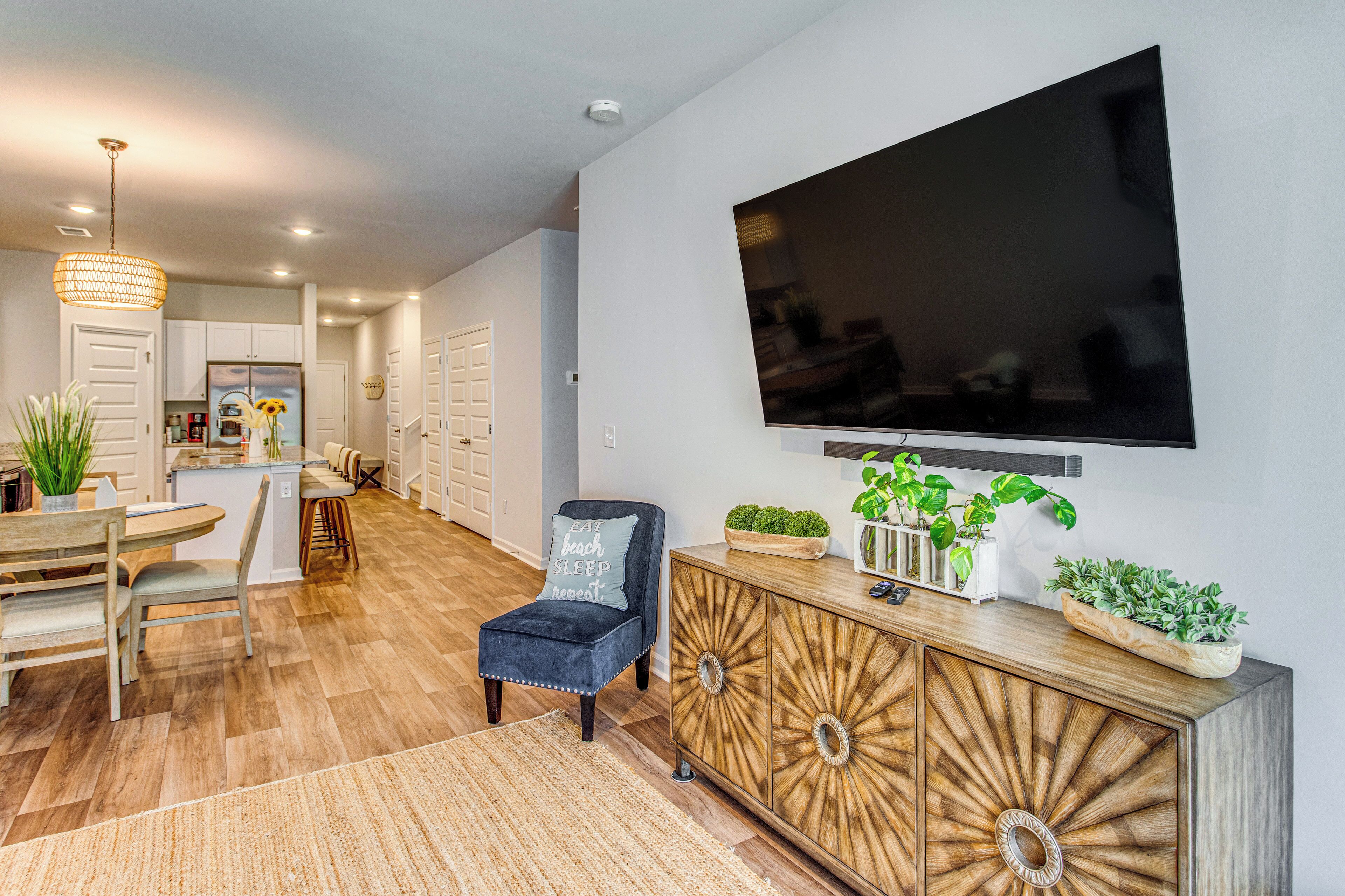 A modern living room with a flat-screen TV, a wooden cabinet with plants, a blue chair, and a dining area with a wooden table and chairs.