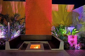 Un'area salotto esterna moderna con un elemento decorativo a fuoco, circondata da piante e pareti illuminate.