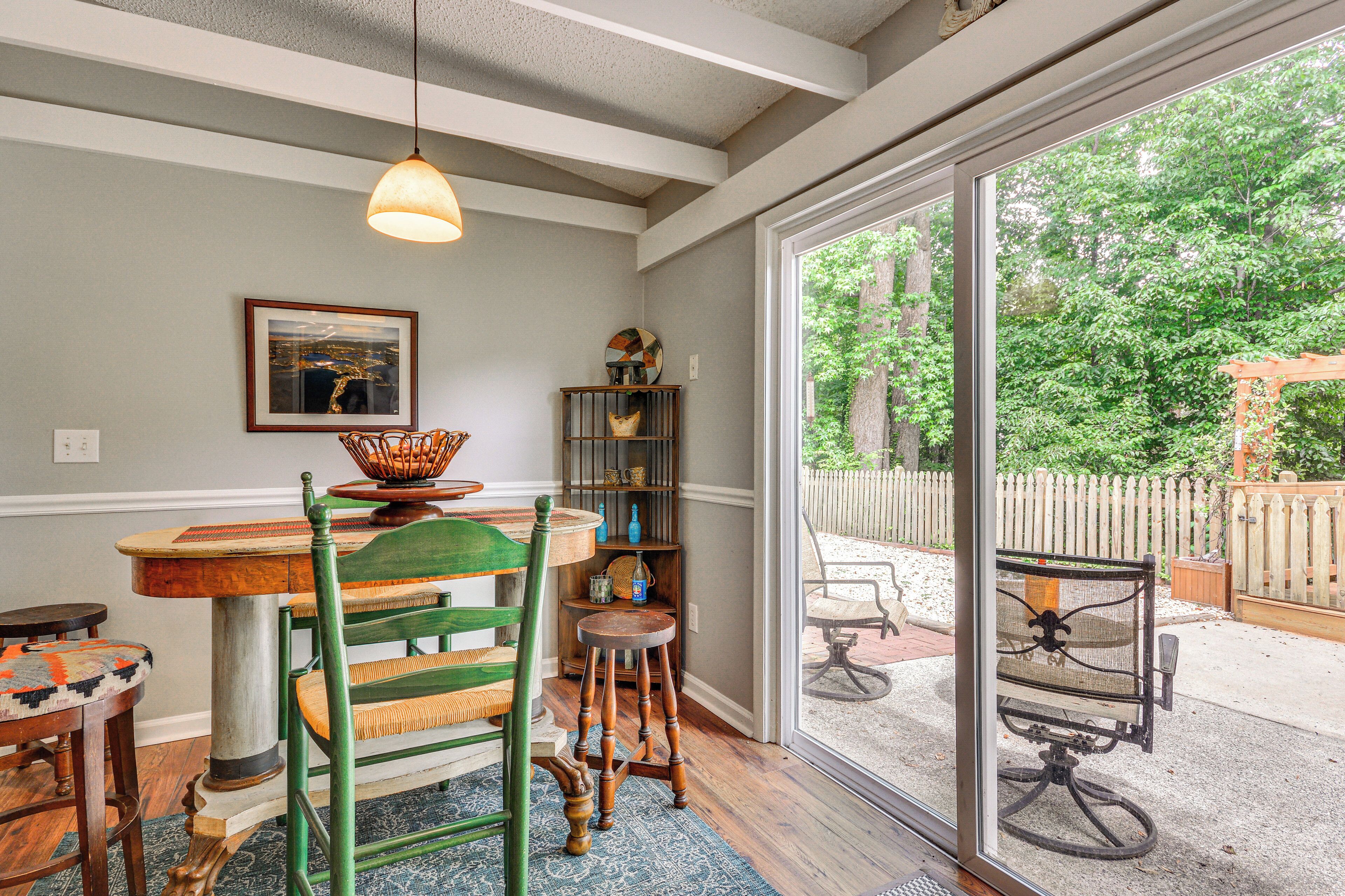 A dining area with a wooden table, chairs, a hanging light fixture, a bookshelf, and a large window with a view of a fenced backyard.