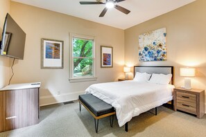 Quarto com cama, mesas de cabeceira, escrivaninha, um banquinho, ventoinha no teto e obras de arte nas paredes.