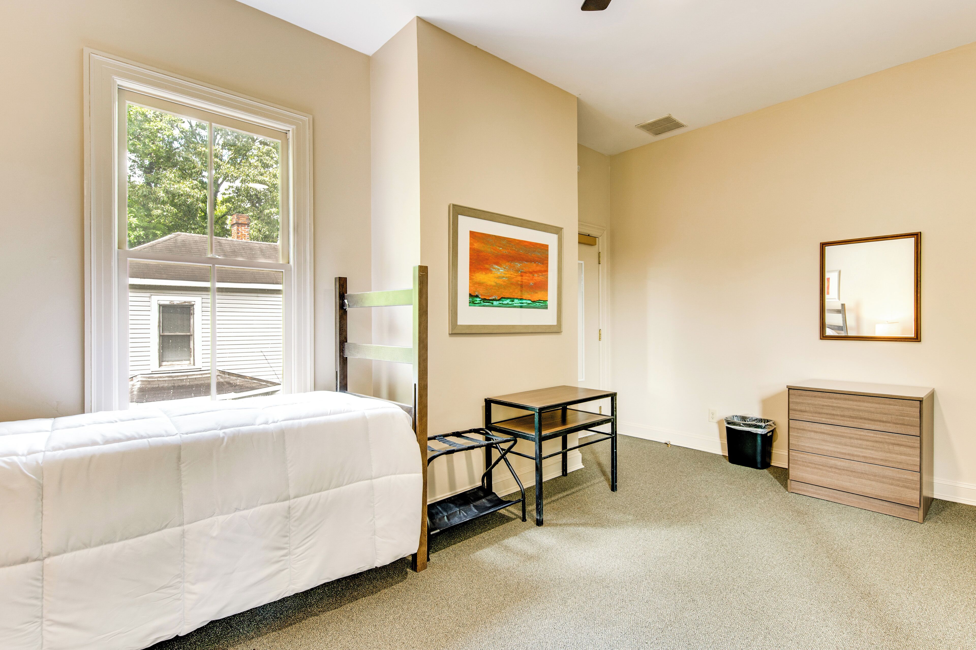 A bedroom with a bed, a desk, a chair, a dresser, and a window with a view of a house.