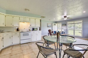 Une cuisine avec des meubles blancs, une table en verre, des chaises en osier et un ventilateur de plafond.