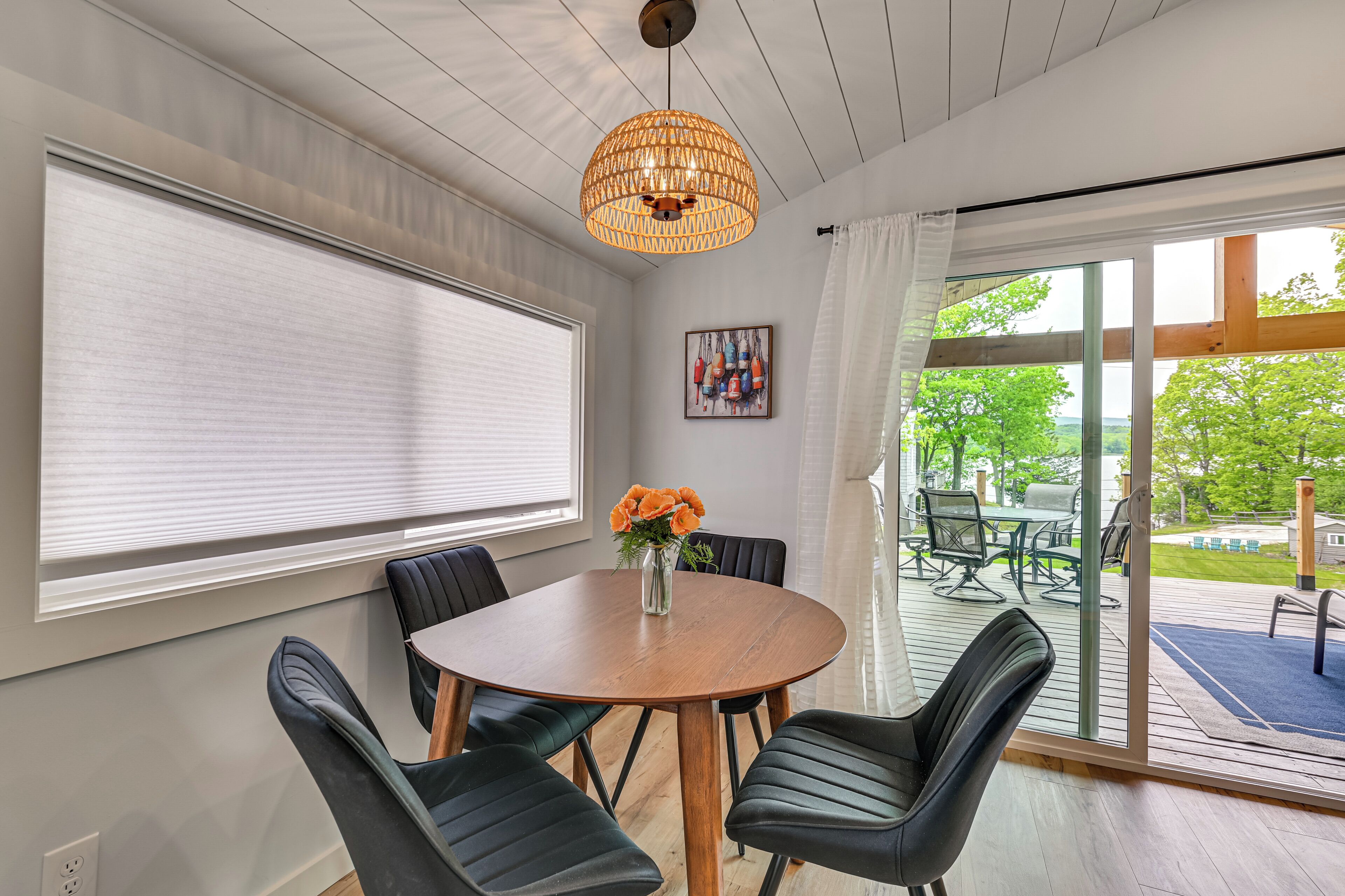 A dining area with a round table, black chairs, a hanging light fixture, a window with blinds, and a view of an outdoor patio.