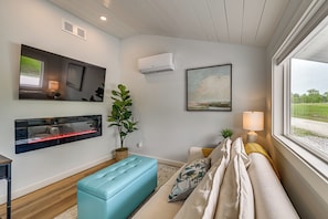 A modern living room with a sofa, a blue ottoman, a wall-mounted TV, an air conditioner, and a window with a view.