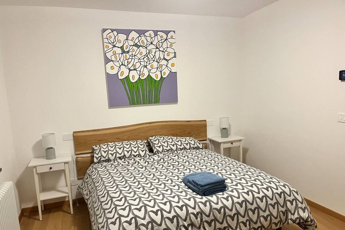 Ein Schlafzimmer mit einem hölzernen Kopfteil, einer gemusterten Bettdecke, zwei Nachttischen mit Lampen und einem Wandbild mit Blumen.