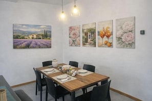 Un coin repas avec une table en bois, des chaises noires et un tableau représentant une maison dans un champ.