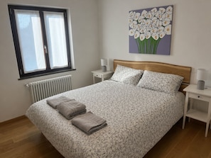 Une chambre à coucher avec un lit, une fenêtre, un radiateur, une table de chevet, une lampe et un tableau.