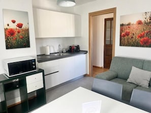 Una cocina moderna con armarios blancos, un microondas y una cafetera. En la zona de estar, hay un sofá verde y una mesa blanca.