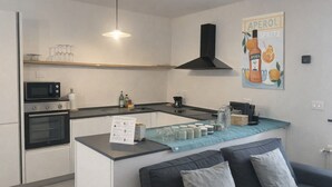 Una cocina moderna con microondas, horno y cafetera. Hay una pequeña isla con un paño azul y frascos. Un sofá con cojines está en primer plano.