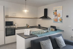 A modern kitchen with a microwave, oven, and a coffee maker. There is a small island with a blue cloth and jars. A sofa with cushions is in the foreground.