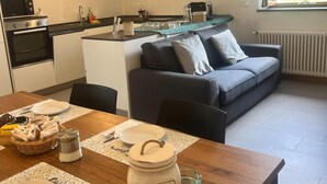 Una cocina funcional con estufa, microondas y cafetera, una mesa de comedor con posavasos, y un sofá gris.